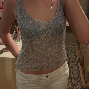 Glittrig silver croppad linnetopp - Säljer en glittrig croppad linnetopp i silver med ribbad struktur. Toppen är ärmlös och har en rundad halsringning, perfekt för att sticka ut på kvällen. Materialet är syntet och ger en skimrande effekt som fångar ljuset snyggt. Aldrig använd