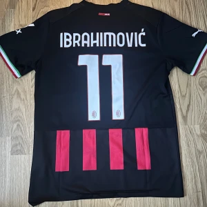 AC Milan Ibrahimović matchtröja Puma - Snygg AC Milan fotbollströja med Ibrahimović och nummer 11 på ryggen. Tröjan är svart med röda vertikala ränder, klubbmärke och Puma-logga på bröstet samt sponsortryck. Ärmarna har italienska flaggans färger vid mudden. Tillverkad i lätt och ventilerande material.