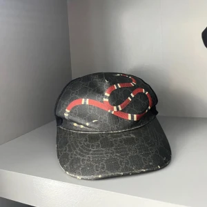 Gucci keps - Snygg svart keps från Gucci med klassiskt GG-monogram och en röd orm som slingrar sig över framsidan. Kepsen har böjd skärm och mesh på baksidan för extra ventilation. Perfekt accessoar för att lyfta din streetwear-look.