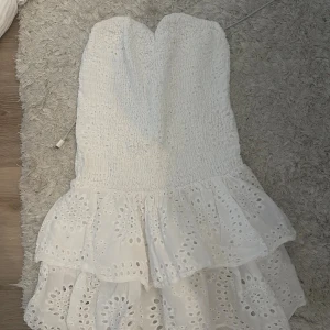 Vit axelbandslös kortklänning med volang - Supersöt vit axelbandslös kortklänning från meet me there med smockad överdel och dubbla volanger i kjolen. Klänningen har broderade detaljer och är perfekt för sommarens alla tillfällen. Tillverkad i bomull för en luftig känsla.