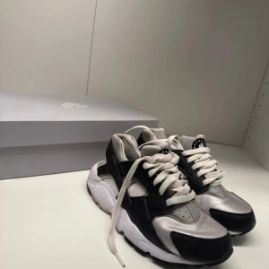 Nike Huarache sneakers svart/vit - Nike Huarache sneakers i svart och vit med silvriga detaljer. Skorna har rund tå, platt sula och snörning framtill. Ovandelen är i syntetmaterial och mesh för extra komfort. Snygg och sportig design med klassisk Huarache-logga på plösen.