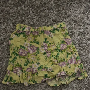 Blommig gul kortkjol från Lindex - Säljer en söt kortkjol från Lindex i storlek 146/152. Kjolen är gul med blommigt mönster i lila, vitt och grönt. Den har volangdetaljer längst ner och resår i midjan för skön passform. Perfekt för dig som gillar färgglada och somriga plagg!