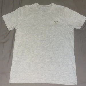 Ljusgrå t-shirt från Prada - Snygg ljusgrå t-shirt från Prada med klassisk passform och korta ärmar. T-shirten har en diskret logga i metall på bröstet och är tillverkad i mjuk bomull. Perfekt för dig som gillar stilrena och exklusiva plagg.