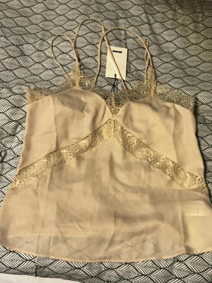 Beige linnetopp med spets  - Säljer en beige linnetopp från  bikbok i storlek S. Toppen har tunna axelband och dekorativa spetsdetaljer längs halsringningen och diagonalt över framsidan. Materialet är mjukt och har en lätt glansig finish som ger en lyxig känsla.