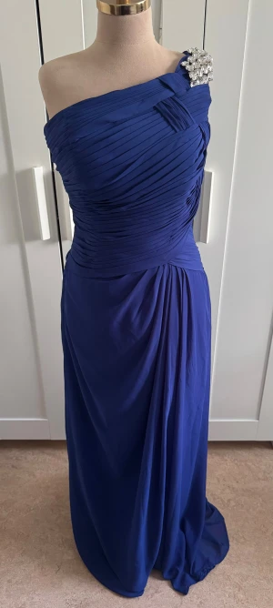 Marinblå långklänning från Izidress - Säljer en marinblå långklänning från Izidress med oneshoulder-design och glittriga stenar på axeln. Klänningen har draperad överdel och mjukt fallande tyg som ger en elegant siluett. Perfekt för bal eller festliga tillfällen.