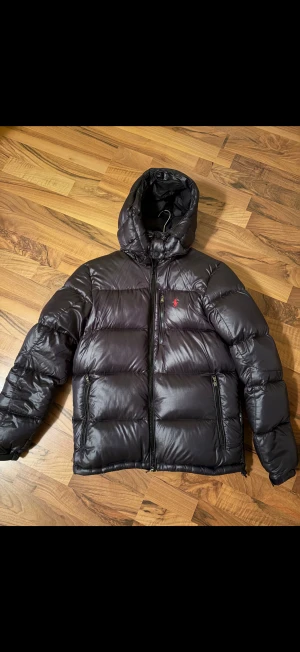 Svart pufferjacka från Polo Ralph Lauren - Svart pufferjacka från Polo Ralph Lauren med huva och två sidofickor med dragkedja. Jackan har en röd Polo-logga på bröstet och quiltad design. Materialet är glansigt och syntetiskt, perfekt för kalla dagar. Stängs med dragkedja framtill. Litet hål på höger arm men syns inte 