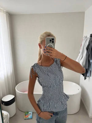 Blårutig blus med knyt på axlarna - Supersöt blus i blåvitt rutigt mönster med knytband på axlarna och volang nedtill. Modellen är figurnära med en smockad midja som ger en snygg siluett. Perfekt till jeans eller kjol för en fräsch look. Materialet känns som bomull och är lätt och luftigt.