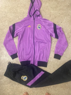 Real Madrid lila och svart träningsset - Säljer ett fett Real Madrid träningsset från Adidas med lila huvtröja och svarta byxor. Jackan har huva, dragkedja och klubbmärke på bröstet, samt svarta detaljer på ärmarna. Byxorna är svarta med lila panel och klubbmärke på benet. Perfekt för dig som älskar fotboll och vill sticka ut.