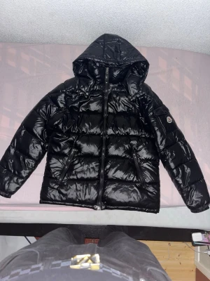 Svart Moncler pufferjacka med huva - Svart glansig pufferjacka från Moncler med huva och dragkedja framtill. Jackan har två sidofickor med dragkedja och Moncler-logga på vänster ärm. Perfekt för kalla dagar och har en riktigt clean look. Priset kan diskuteras. (Hör av dig för mer bilder)