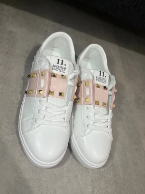 Valentino Rockstud Untitled sneakers - Säljer ett par vita Valentino Rockstud Untitled sneakers med ljusrosa band och guldiga nitar över ovansidan. Skorna har snörning, rund tå och platt sula. Materialet är skinn och detaljerna ger en lyxig vibe. Kommer med originalkartong och dustbag. Passar dig som vill ha en lyxig stil.