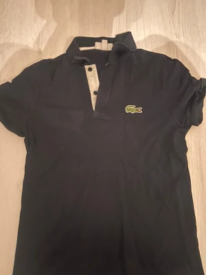 Svart pikétröja från Lacoste - Klassisk svart pikétröja från Lacoste med vit knappslå och det ikoniska gröna krokodilmärket på bröstet. Tröjan har korta ärmar, krage och knappar framtill. Perfekt för en stilren och sportig look. Små pytte hål längst ner vid magen då katten råka dra lite i den men inget märkbart enligt mig 