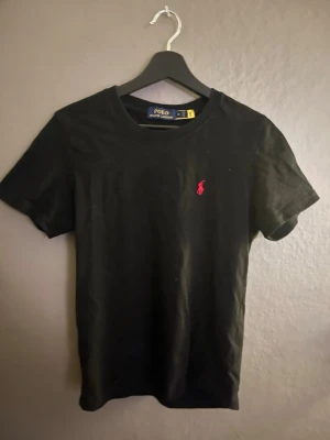 Svart t-shirt från Polo Ralph Lauren - Svart t-shirt från Polo Ralph Lauren med klassisk passform och korta ärmar. Ikoniska röda loggan broderad på bröstet. Tillverkad i mjuk bomull som känns skön mot huden. Perfekt till jeans eller shorts för en clean och stilren look.