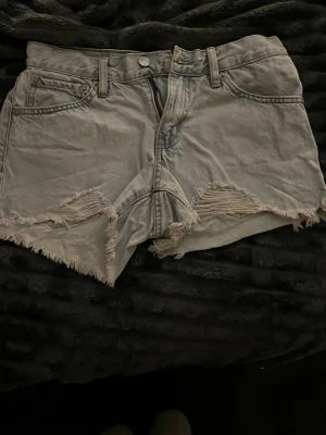 Ljusgrå denimshorts med fransad kant - Säljer ett par ljusgrå denimshorts från 157 med fransad kant och slitna detaljer framtill. Klassisk femficksmodell med bälteshällor och knappstängning. Perfekta för varma dagar och har en cool, avslappnad vibe.