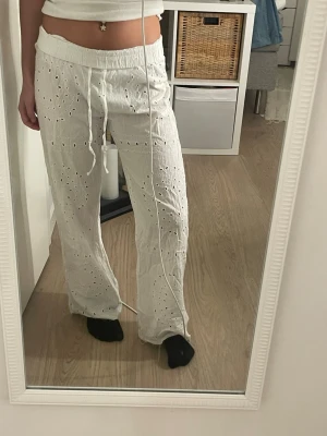 Vita broderade byxor från Pull&Bear - Säljer ett par vita byxor från Pull&Bear i storlek XS. Byxorna har broderade hålmönster, elastisk midja med snörning och är tillverkade i ett lätt bomullsmaterial. Passformen är loose och modellen är rak med avslappnad stil.