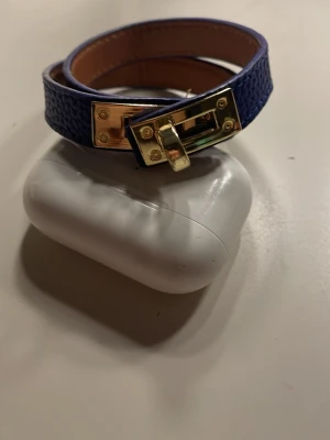 Blå skinnarmband med gulddetaljer - Snyggt blått armband i skinnimitation med guldfärgat spänne och struktur på ytan. Armbandet har en elegant låsdetalj i metall och är fodrat med ljusbrunt material på insidan. Perfekt accessoar för att lyfta din outfit.