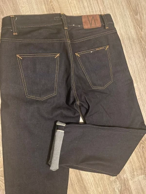 Selvedge jeans från Nudie - Säljer ett par mörkblå jeans från Nudie Jeans med klassisk orange kontrastsöm och läderpatch bak. Modellen har raka ben och normal passform. Jeansen är tillverkade i kraftig denim av bomull och har snygg selvedge-detalj vid uppviket. Nypris ligger på 2000kr, pris kan diskuteras!