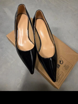 Svarta pumps med spetsig tå och låg klack - Snygga svarta pumps med glansig finish och spetsig tå. Skorna har låg klack och en klassisk, elegant form. Klackens höjd 3cm. Insidan är beige och materialet ser ut att vara syntet med lackad yta. Perfekt för dig som gillar stilrena och tidlösa skor.