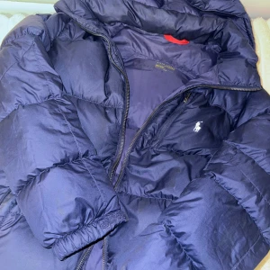 Mörkblå pufferjacka från Polo Ralph Lauren - Säljer en mörkblå pufferjacka från Polo Ralph Lauren med klassisk logga på bröstet. Jackan har hög krage, dragkedja och är tillverkad i polyester. Perfekt för kalla dagar och har en schysst oversized vibe.