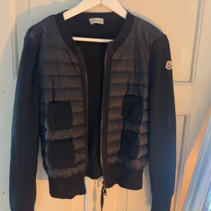 Svart quiltad cardigan från Moncler - Svart pufferjacka från Moncler med quiltad front och stickade ärmar. Jackan har två stora fickor fram, dragkedja och Moncler-logga på ärmen. Materialmix av polyester och stickad ull ger en schysst kontrast. Perfekt för dig som gillar stilren och sportig vibe.