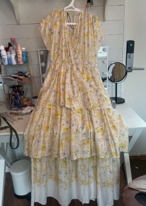 Blommig långklänning från H&M - En ljus och romantisk klänning med gult blommönster, v-ringning och mjuka, korta ärmar. Den har markerad midja och en luftig, lager-på-lager-kjol som ger ett elegant och somrigt fall. Jag hade på mig den på min kusins konfirmation