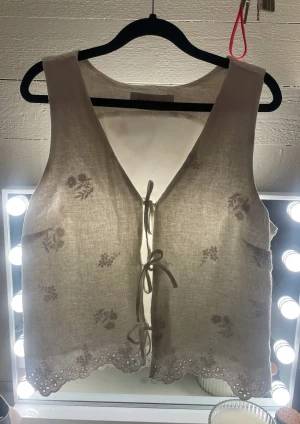 Vit blommig väst från Hollister - Säljer en söt vit väst från Hollister med broderade blommor och knytning framtill. Västen har v-ringad hals och spetsdetaljer längs nederkanten. Materialet är linne och ger en luftig känsla, perfekt för varmare dagar.