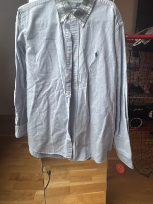 Blå skjorta från Polo Ralph Lauren - Säljer en blå skjorta från Polo Ralph Lauren i storlek L. Skjortan har en rutig krage i grönt, blått och gult, klassisk knappstängning och lång ärm. Materialet är mjuk bomull och den har den ikoniska logon broderad på bröstet.