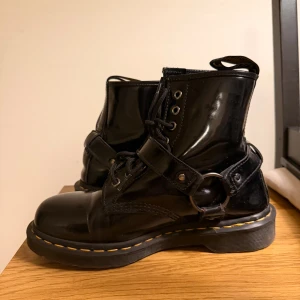 Svarta Dr. Martens boots i skinn - Säljer ett par svarta Dr. Martens boots i blankt skinn med klassisk gul söm runt sulan. Skorna har snörning, spänne och metallring som cool detalj. Grov platt sula och dragflik med AirWair-tryck. Perfekta för dig som vill sticka ut med din stil. Köpta för 2000kr.