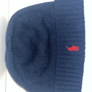 Mörkblå stickad mössa från Polo Ralph Lauren - Mörkblå stickad mössa från Polo Ralph Lauren med röd broderad logga framtill. Klassisk ribbad kant och mjukt material som håller dig varm under kalla dagar. Perfekt accessoar för en stilren look.