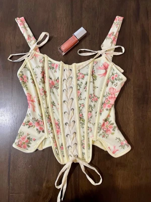 Blommig korsett med snörning - Superfin korsett i ljusgul med rosa blommor och rosett-detaljer. Toppen har snörning framtill, axelband med knyt och markerade sömmar för en snygg siluett. Materialet känns som polyester och har en mjuk, lätt struktur. Perfekt för dig som gillar romantiska och söta vibes.