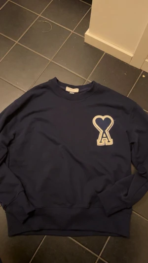 Mörkblå sweatshirt från Ami Paris - Snygg mörkblå sweatshirt från Ami Paris med stort broderat hjärta och bokstaven A på bröstet i beige. Tröjan har rund hals, långa ärmar och ribbade muddar. Perfekt för dig som gillar stilrena och trendiga plagg med en twist.