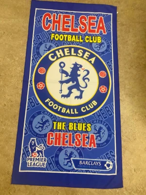 Chelsea FC blå fotbollsväst - Säljer en blå väst med Chelsea Football Club-tryck och Premier League-logga. Västen har ett stort lejonemblem i mitten och texten 'THE BLUES CHELSEA' i rött och gult. Materialet är troligtvis polyester och designen är sportig med tydliga klubbfärger.