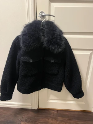 Marin blå teddyjacka med päls från Gina Tricot - Svart teddyjacka från Gina Tricot med fluffig pälsimitation på kragen. Jackan har två stora fickor med knappar framtill och en croppad, boxig passform. Materialet är mjukt och strukturerat, perfekt för dig som gillar en cool och mysig look.