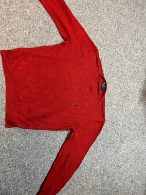 Röd stickad tröja från Polo Ralph Lauren - Säljer en röd stickad tröja från Polo Ralph Lauren med klassisk blå broderad logga på bröstet. Tröjan är tillverkad i 100% bomull, har rund hals och långa ärmar med ribbade muddar vid ärmslut och nederkant. Perfekt för dig som gillar stilren design.äkta
