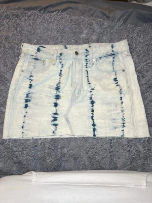 Vit och blå kortkjol från & Denim - Säljer en kort jeanskjol från & Denim med coolt batikmönster i vitt och blått. Kjolen har klassiska fem fickor, knappgylf och rå kant nedtill för en avslappnad vibe. Perfekt för dig som gillar streetstyle och vill sticka ut lite extra.
