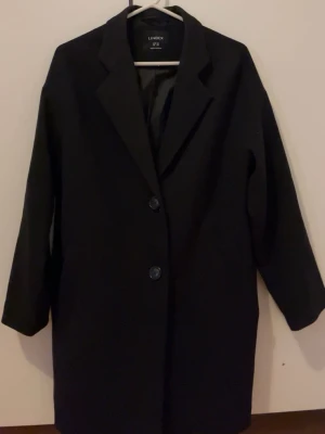Svart kappa från Lindex XS - Stilren svart kappa från Lindex i storlek XS. Klassisk rak modell med krage och två knappar framtill. Långärmad och med slät yta, perfekt för dig som gillar enkel och snygg design. Passar dig som vill ha en tidlös look.