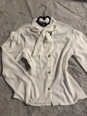 Vit blus med rosett  - Snygg vit blus med knytrosett i halsen och dekorativa knappar framtill. Perfekt för dig som gillar stilrena detaljer. Helt ny 