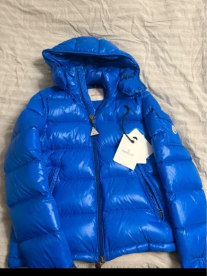Blå Moncler dunjacka med huva - Säljer en fet blå dunjacka från Moncler med huva och coolt Moncler-märke på ärmen. Jackan har dragkedja, sidofickor och ett unikt serietidningsfoder på insidan. Materialet är glansigt och känns lyxigt, perfekt för kalla dagar. MÖTS BARA ‼️