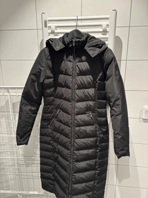 Svart lång dunjacka från Rockandblue - Otrolig dunkappa från Rockandblue med fina detaljer. Väl omhändertagen, det enda att anmärka på är pyttelite smink på kragen som inte syns när jackan är på, men går säkert att få bort. Figursydd! Jag är S i vanliga fall och den passar perfekt