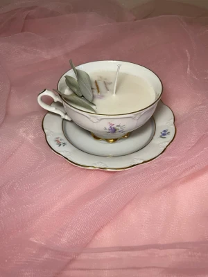 Doftljus i en tekopp i en vit färg med blommig design  - Det här ljuset är gjort av återanvändbara material. Tekoppen är köpt secondhand och stearinet är från kyrkor som tänkte slänga det. Det här ljuset har en svag doft utav vanilj, så känsliga även kan använda det. Det här ljuset är även dekorerat med torkade blad och kanel 