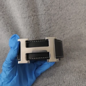 Svart skärp med H-spänne i silver - Snyggt svart skärp med ett stort H-format spänne i silverfärgad metall. Skärpet har vita sömmar längs kanterna och är tillverkat i slätt skinn. Perfekt för att ge outfiten en lyxig touch och passar till jeans eller kostymbyxor.