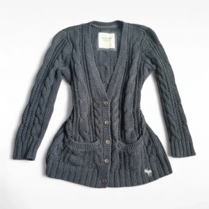 SVART stickad kofta Abercrombie - OBS! TRÖJAN ÄR SVART INTE GRÅ!!! Snygg stickad kofta från Abercrombie & Fitch med grova flätstickade detaljer, v-ringning och knappar framtill. Koftan har två fickor och en liten broderad logga vid nederkanten. Materialet är bomull och akryl, perfekt för lager på lager under kyliga dagar, pris kan diskuteras!!