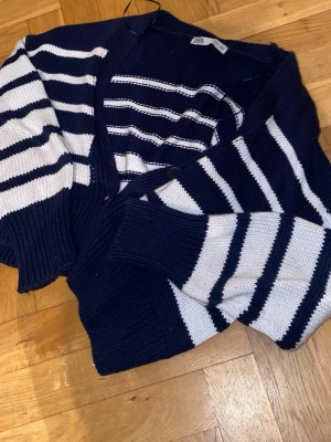 Marinblå och vit randig kofta från Zara - Säljer en snygg croppad stickad kofta från Zara i storlek L. Koftan har breda marinblå och vita ränder, långa ärmar och knappar framtill. Perfekt för dig som gillar klassisk stil med en modern twist. Materialet är mjukt och stickat för extra komfort.