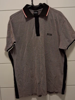 Randig polotopp från Hugo Boss - Pikétröja från Hugo Boss med diskret randigt mönster i svart,har svart krage med röda och vita detaljer, korta ärmar och knäppning framtill. Boss-logga broderad på bröstet.