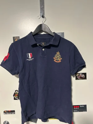 Morris mörkblå pikétröja med brodyr - Mörkblå pikétröja från Morris med broderade emblem och text på bröstet samt en stor röd 68 på ärmen. Klassisk krage och två knappar framtill. Tröjan är i bomull och har en sportig, stilren look.