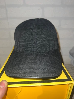 Svart Fendi keps med mönster - Snygg svart keps från Fendi med diskret ton-i-ton mönster och justerbar rem i skinn baktill. Kepsen har klassisk form och är tillverkad i ett slitstarkt material som ger en lyxig känsla. Perfekt för dig som vill ha en stilren accessoar med designer-vibe.