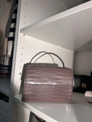 Ljubrun handväska i croco-imitation - Snygg ljusbrun  handväska med croco-mönstrad yta och stilren, boxig form. Väskan har ett topphandtag och medföljande axelrem, perfekt för att lyfta vilken outfit som helst. Magnetlås framtill och glansig finish ger en trendig vibe.