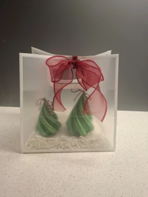 Grön julgran-väst i stearin - Unik väst formad som en julgran i grön stearin med skimrande detaljer och snurrad design. Toppen har en liten rosett i rött och vitt snöre, perfekt som dekor till jul. Säljs i set om två, levereras i fin presentförpackning med röd rosett.