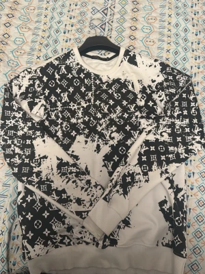 Louis Vuitton svartvit sweatshirt - Svartvit sweatshirt från Louis Vuitton med ikoniskt LV-mönster över hela tröjan. Tröjan har långärmad design, rund hals och ribbade muddar vid ärmslut och nederkant. Materialet känns mjukt och bekvämt, perfekt för dig som gillar streetstyle.