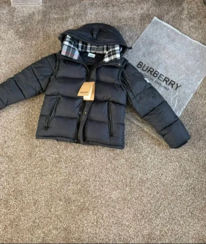Svart pufferjacka från Burberry - Svart pufferjacka från Burberry med avtagbara ärmar, huva och klassiskt rutigt foder i svart, vitt och rött. Jackan har dragkedja framtill, två sidofickor med dragkedja och en snygg, boxig passform. Perfekt för kalla dagar och har en modern vibe.