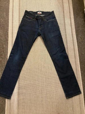 Tommy Jeans Slim Scanton blå jeans - Snygga mörkblå jeans från Tommy Jeans, modell Slim Scanton. Klassisk femficksdesign med gul kontrastsöm och en liten ficka med Tommy Hilfiger-flagga. Slim fit och raka ben, tillverkade i jeansmaterial. Perfekt för dig som gillar stilrena och moderna jeans.